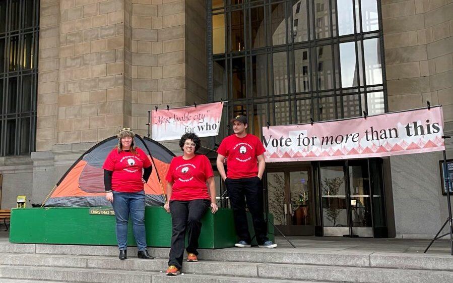 Three Pittsburgh DSA volunteers stand in front of Pittsburgh's City County Building to ask "most livable city...for who?" and to demand representatives "vote for more than this." The status quo is represented by a camping tent, which is all some of Pittsburghers have for shelter. Photo by Greg Sinn