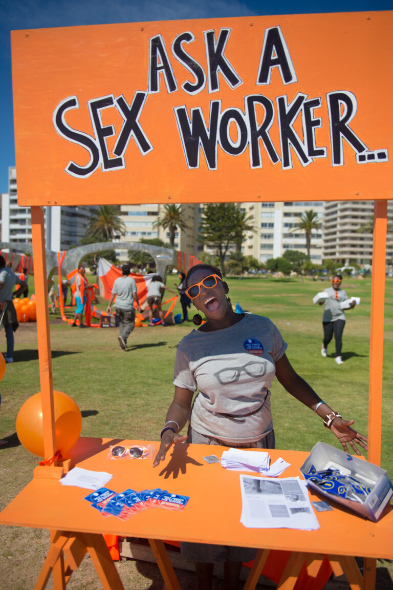 A person standing an orange stand with a sign above reading, "ask a sex worker"