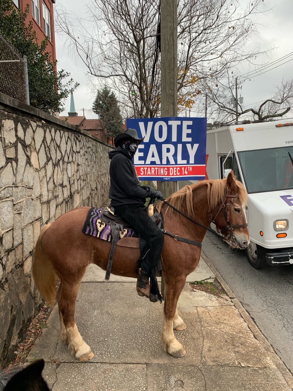 PHOTO from unstoppable voters, cowboy in all black on. abrown horse on a side walk in an urban area, with a large Vote Early sign